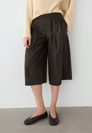 Person iført løstsiddende sorte knælange culottes, beige langærmet top og sorte flade sko, stående på en grå gulv mod en hvid væg.