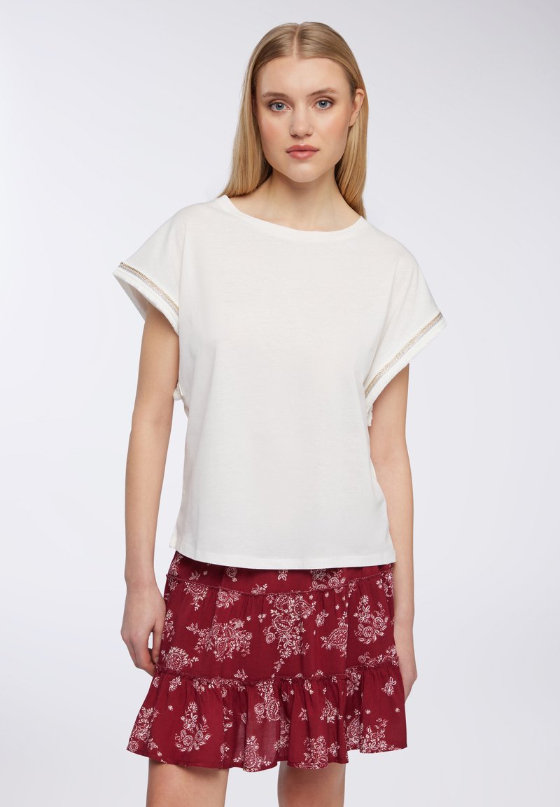 Top bianco a maniche corte con scollatura ampia, caratterizzato da cuciture decorative dorate sulle maniche. Abbinato a una gonna ruffled a stampa floreale borgogna.