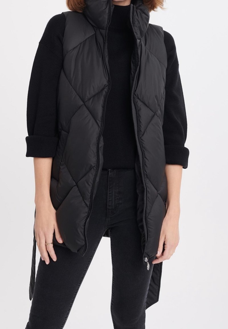 Femme portant une veste sans manches matelassée noire sur un pull noir à manches longues et un jean noir, les mains détendues le long du corps.