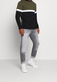 Sudadera negra y verde oliva con una franja horizontal blanca, combinada con jeans gris claro desgastados y zapatillas altas blancas.