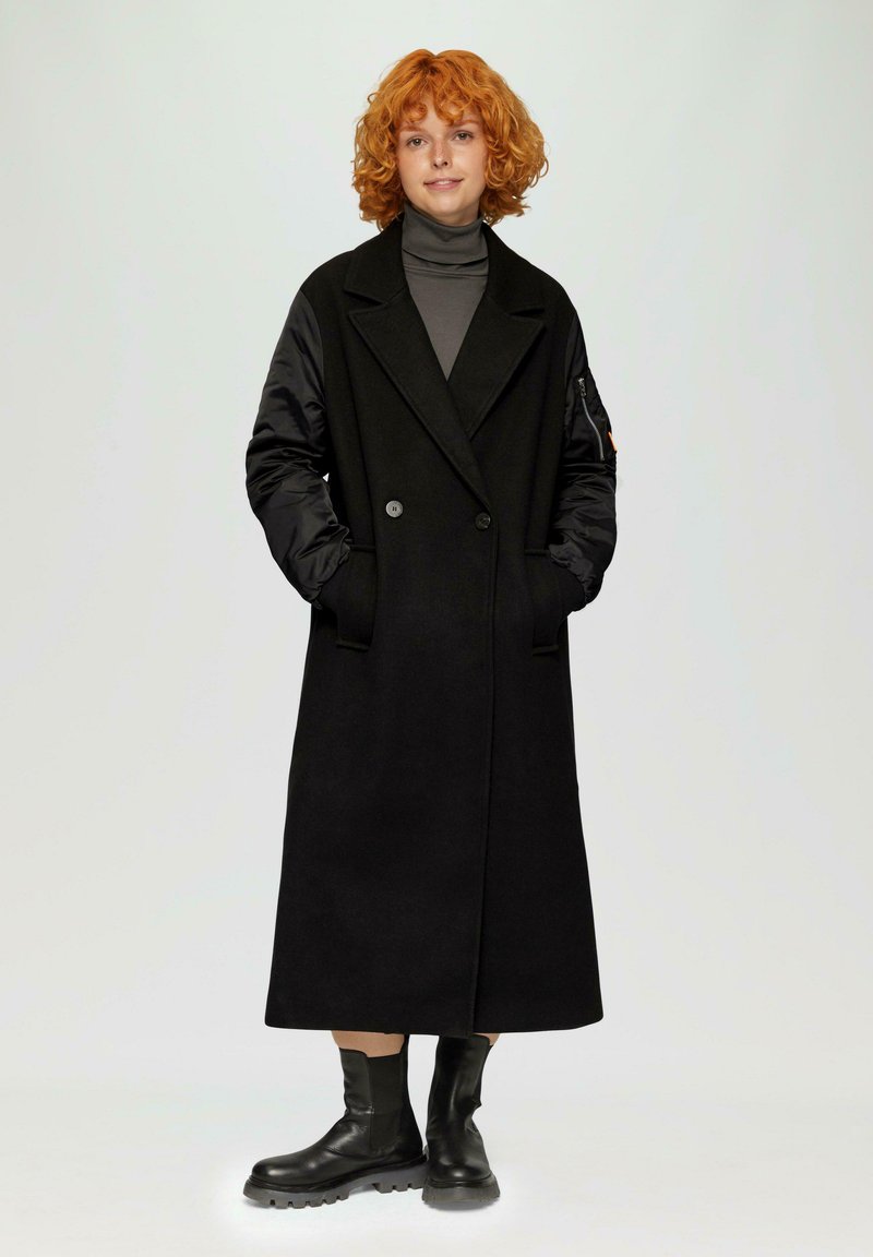 Lang zwarte jas met contrasterende zwarte gewatteerde mouwen, dubbelzijdige voorkant en zijzakken. G gedragen over een grijze coltrui. Zwarte laarzen.