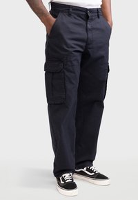 Schwarze Cargo-Hosen aus Baumwolle mit großen Seitentaschen, lockerer Passform und Reißverschluss, kombiniert mit schwarz-weißen Sneakers.