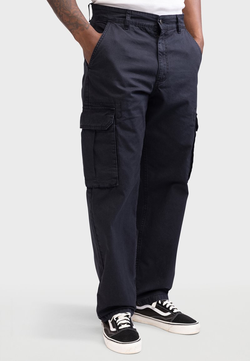 Schwarze Cargo-Hosen aus Baumwolle mit großen Seitentaschen, lockerer Passform und Reißverschluss, kombiniert mit schwarz-weißen Sneakers.