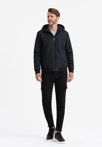Svart huva jacka med framzip, två stora fickor och ribbad mudd, i kombination med svarta cargobyxor och svarta sneakers med vita sulor.