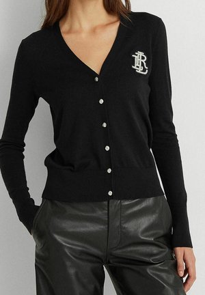 Femme portant un cardigan noir boutonné avec un logo blanc sur la poitrine et un pantalon en cuir noir, les mains dans les poches.