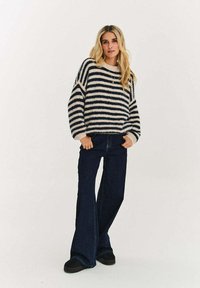 Sweter w paski w kolorze kremowym i granatowym, oversize z szerokimi rękawami, zestawiony z ciemnymi jeansami o rozszerzanych nogawkach i czarnymi butami, na neutralnym tle.