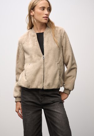 Giacca bomber in pelliccia sintetica beige con zip frontale, due tasche laterali, polsini a coste e vestibilità comoda, indossata sopra un top nero e jeans scuri.