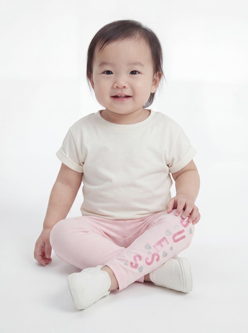 Tout-petit assis en tailleur portant une chemise blanche, un pantalon rose avec texte pailleté et des chaussures blanches, souriant à la caméra sur fond blanc.
