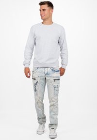 Felpa grigia con polsini a costine, abbinata a jeans di denim azzurro chiaro con cuciture decorative e diverse tasche. Sneakers bianche completano il look.