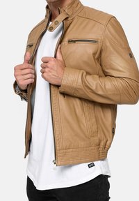 Hellbraune Lederjacke mit Stehkragen, Reißverschlüssen vorne und gerippten Akzenten an Schultern und Bündchen. Gefüttert mit gestreiftem Muster.