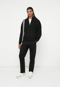 Maglione nero a maglia con design a mezze cerniera, abbinato a pantaloni cargo neri. Gli accessori includono una borsa a tracolla beige e sneakers bianche.