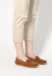 Braune Wildleder-Loafers mit gestickten Details, flachen Gummisohlen und abgerundeter Zehenform, kombiniert mit beigefarbenen, knöchellangen Hosen.