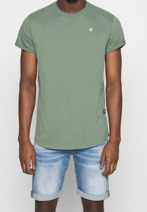 Homme portant un t-shirt vert olive à manches retroussées associé à un short en jean bleu clair retroussé au-dessus des genoux.