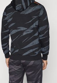 Zwarte hoodie met grijze abstracte patronen, voorzien van een kangoeroezak en een geribbelde onderrand. Ronde capuchon met een zichtbaar logo op de schouder.