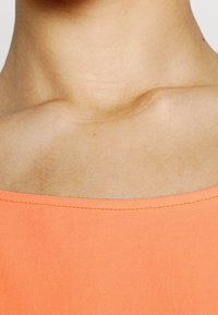 Růžovo-oranžový top s širokým výstřihem a viditelnými švy, kontrastující s hladkou pletí pod klíční kostí.