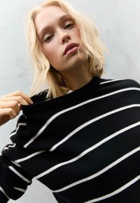 Maglione nero con strisce orizzontali bianche. Materiale a maglia, scollatura ampia e polsini a coste. Il modello tocca il colletto con una mano.