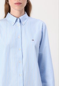 Ljust blå skjorta med knappar, vertikala vita ränder, spetsig krage, långa ärmar och en liten broderad logotyp på bröstet.
