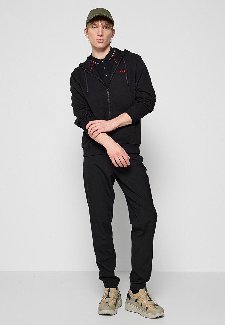 Schwarze Zip-Hoodie mit roten Akzenten, kombiniert mit schwarzen Hosen und beigen Sneakers. Das Modell trägt eine grüne Mütze und steht vor einem neutralen Hintergrund.