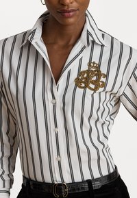 Cămașă în dungi cu linii verticale negre și albe, având un inscripționat auriu brodat pe partea stângă a pieptului și butoane albe.