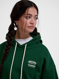 Ung kvinde med langt flet hår iført sølv hoop øreringe og en mørkegrøn Hummel sportswear hættetrøje, der kigger mod højre.