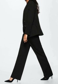 Blazer noir ajusté avec des revers pointus, associé à un pantalon droit noir taillé sur mesure. Porté avec des chaussures noires à talons hauts. Texture de tissu lisse.