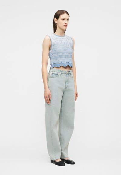 Νεαρή γυναίκα που στέκεται φορώντας ένα αμάνικο crop top με μπλε κυματιστό σχέδιο, ανοιχτόχρωμα τζιν παντελόνια φαρδιάς γραμμής και μαύρες μπαλαρίνες, μπροστά σε λευκό φόντο.