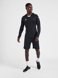 Zwart sportshirt met lange mouwen en witte logo's en schouderaccenten, gecombineerd met zwarte shorts en zwarte sportschoenen met witte zolen.