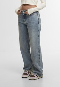 Osoba ubrana w jasnoniebieskie szerokie dżinsy z postrzępionymi nogawkami oraz dwukolorowe trampki, poprawiająca pas dżinsów na tle jednolitego tła.