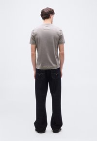 Άνδρας που στέκεται πλάτη φορά γκρι κοντομάνικο πουκάμισο και σκούρο μπλε τζιν μπροστά σε απλό λευκό φόντο.