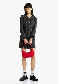 Abito in denim grigio scuro, cinture in vita, abbinato a una borsa in nylon rossa. Il modello indossa calze bianche con strisce nere e rosse e scarpe nere.