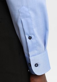 Camicia a maniche lunghe azzurro chiaro mostrata, con polsino abbottonato con due bottoni scuri e una texture liscia. Tessuto scuro visibile parzialmente.