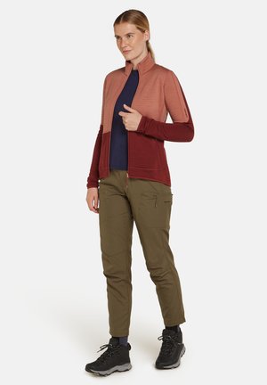 Rode en roze zip-up jas met lange mouwen, textuurstof. Gecombineerd met olijfgroene broek met ritszakken en zwarte wandelschoenen.