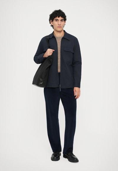 Man stands wearing navy jacket, brown sweater, dark corduroy pants, black shoes, holding large black shoulder bag against plain background.