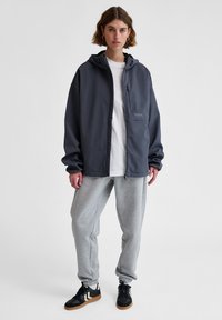 Mörkgrå zip-up jacka med huva, har en bröstficka och kontrasterande sömmar, tillsammans med ljusgrå joggers och svarta sneakers.