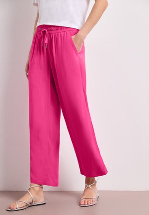 Femme portant un pantalon large rose vif avec une ceinture à cordon, un haut blanc et des sandales plates argentées à brides, debout contre un mur clair.