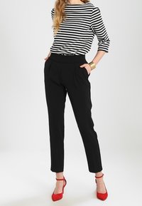 Woman in black high-waist trousers, black-and-white striped 3/4 sleeve top, red ankle-strap heels, gold cuff bracelet, hands in pockets.