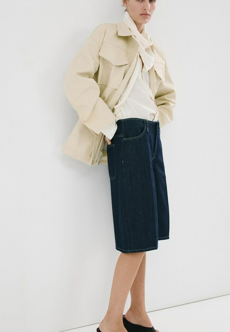 Personne portant une veste beige, un chemisier blanc à col noué, un short en jean bleu foncé à hauteur des genoux et des chaussures noires sans lacets, debout contre un mur blanc.