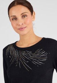 Suéter negro con un escote redondeado y elaborados adornos de strass en plata y oro dispuestos en un patrón fluido.