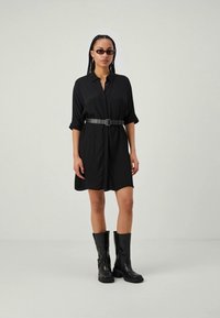 Schwarzes Hemdkleid mit lockerer Passform, umgeschlagenen Ärmeln und einem taillierten Bund. Getragen mit knielangen schwarzen Stiefeln und Sonnenbrille. Glatte Stofftextur.