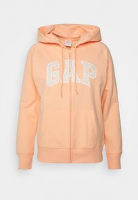 Světlý broskvový zipový svetr s předními kapsami, kapucí na šňůrku a velkým bílým nápisem "GAP" přes hrudník.