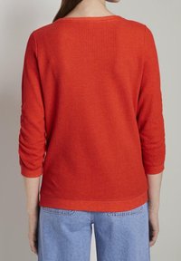 Top largo de manga larga rojo, con canalé, cuello redondo y puños ajustados, combinado con jeans azul claro, mostrando la vista trasera.