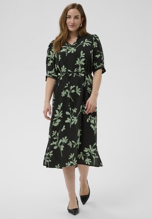 Woman wearing a black midi dress with light green leaf patterns, puffed short sleeves, tied waist, and black pointed-toe heels, standing on white background.