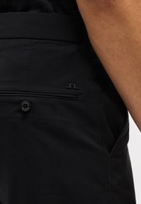 Τσέπη μαύρου παντελονιού με κουμπί και μικρό λογότυπο της μάρκας, που δείχνει μέρος του μαυρισμένου χεριού ενός ατόμου δίπλα στη μέση.