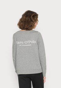 Sudadera gris con un ajuste relajado, con un cuello redondo y el texto "Marc O’Polo EST. EN ESTOCOLMO" impreso en blanco en la parte posterior.