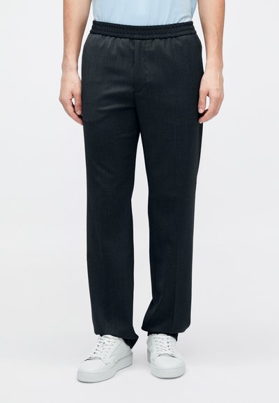 Tumehallid, lõdvalt istuvad püksid elastse vöökohaga, mis on sileda tekstuuriga ja kitsenevate säärtega, kombineeritud valgete tossudega.