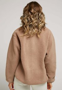 Veste en polaire marron avec une coupe décontractée, surface texturée, épaules droites et ourlet arrondi. Matériau doux et design décontracté.