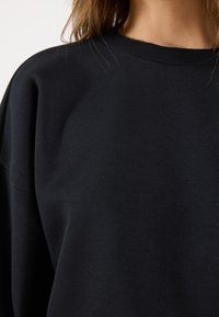 Felpa nera con vestibilità relaxed, scollo a girocollo a costine e texture in cotone morbido. Presenta spalle leggermente scese per un comfort maggiore.