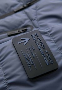 Jasnoniebieska pikowana tkanina kurtki z matową czarną metką z wytłoczonym napisem i wzorem strzałki, przymocowaną blisko zamka.