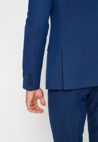 Giacca da abito blu navy con tessuto testurizzato, fessura singola sul retro e quattro bottoni neri sul polsino, che mostrano un design ben aderente.