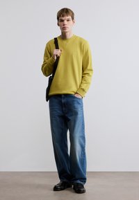 Κίτρινο φούτερ με στρογγυλή λαιμόκοψη και ριμπ μανσέτες, σε συνδυασμό με φαρδιά μπλε τζιν παντελόνια και μαύρα παπούτσια. Απλός σχεδιασμός χωρίς ορατά σχέδια.